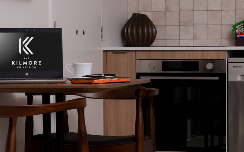 Dining table, with a laptop coffee cup and notebook in the foreground, and the kitchen in the background of the downstairs living area in a one-bedroom self-contained apartment in The Kilmore Collection, Christchurch